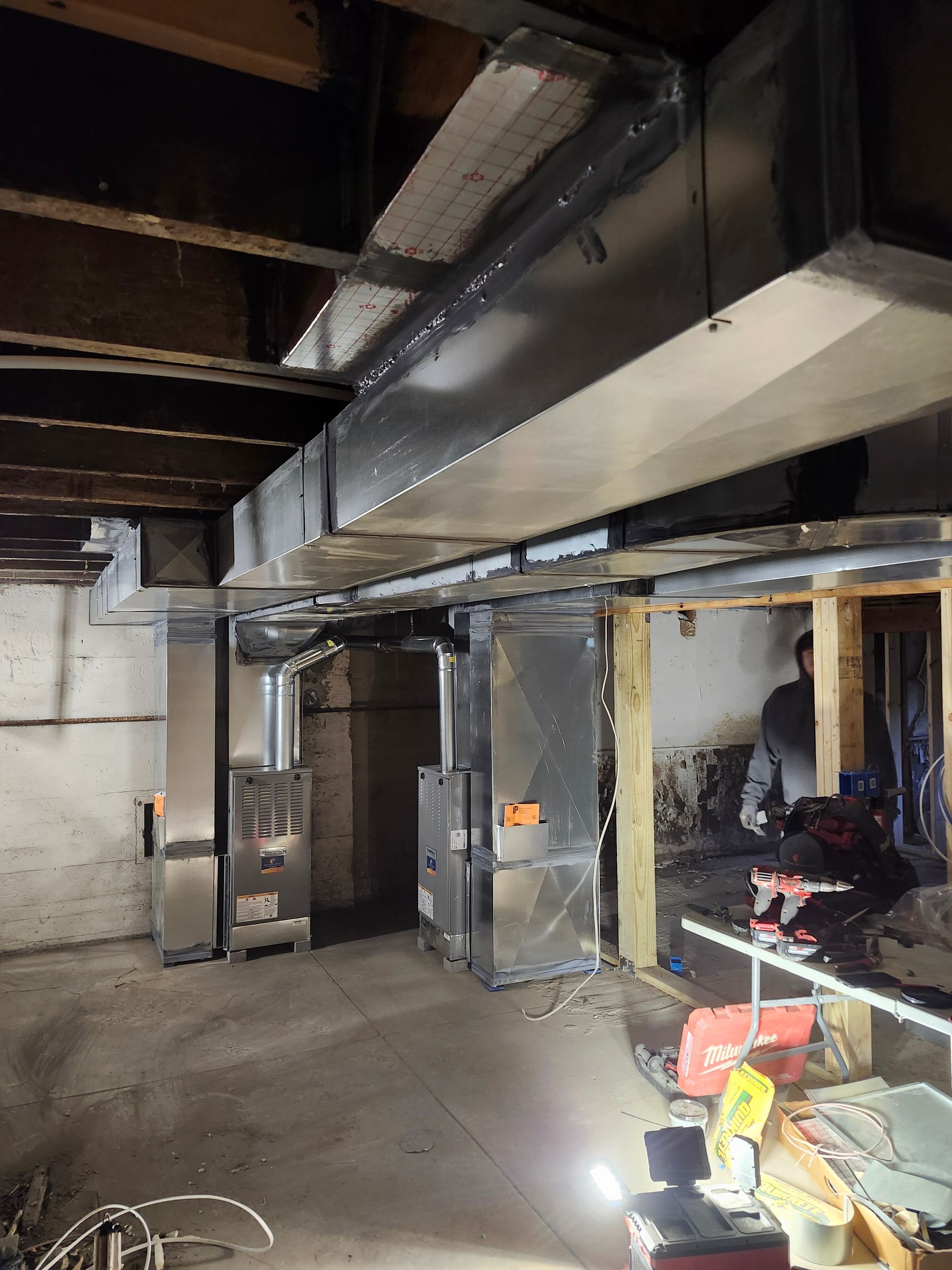 HVAC system installation in a basement. Ductwork and units are visible, along with some tools and a person working.