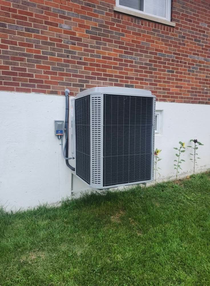 AC unit mounted on white wall, brick building background, green grass below.