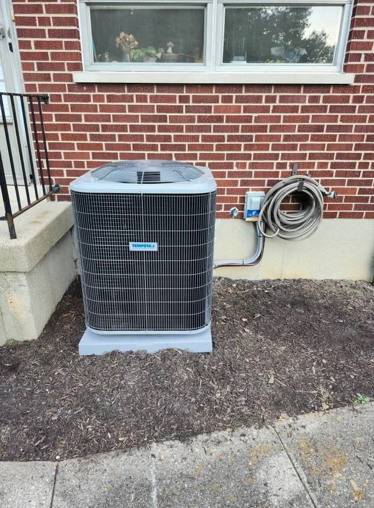 An air conditioning unit against a red brick wall, next to a coiled hose and electrical box.