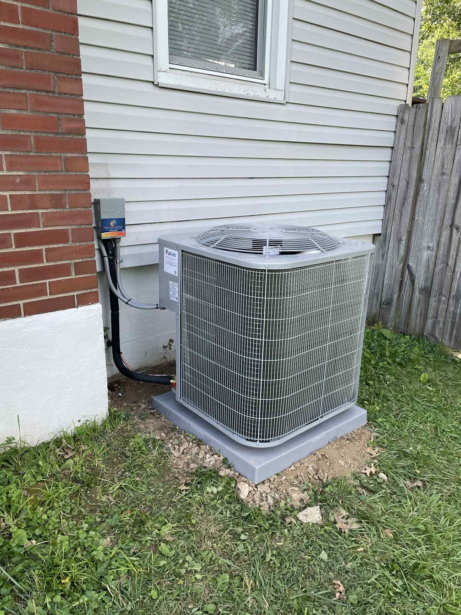 Central air conditioning unit on a concrete pad next to a house with brick and siding, in a grassy yard.