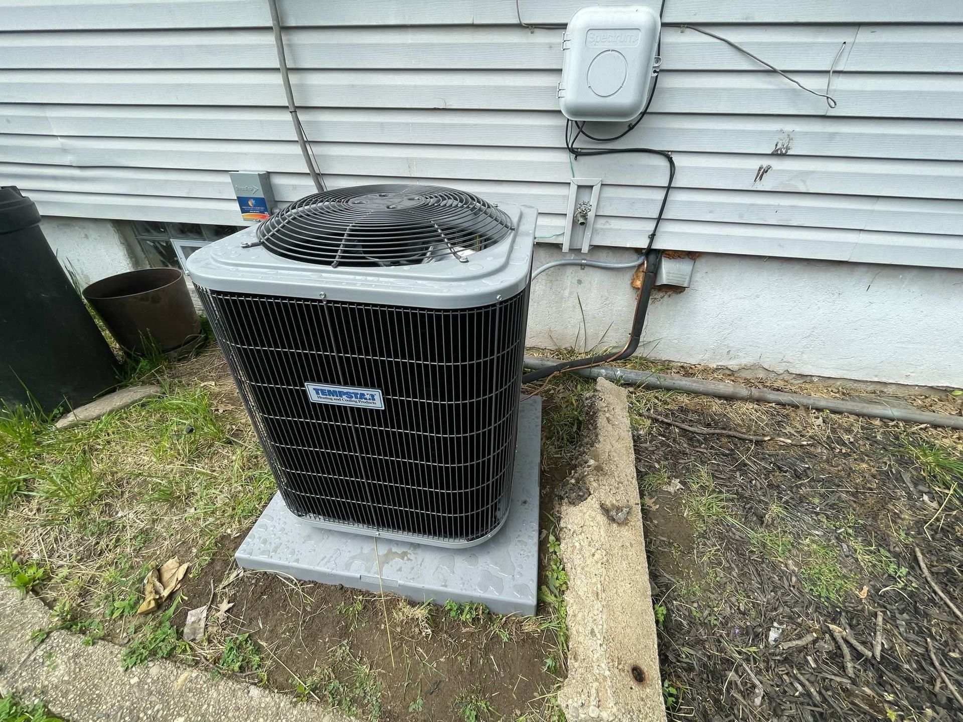 Outdoor air conditioning unit with gray top and black metal fins, beside a house.