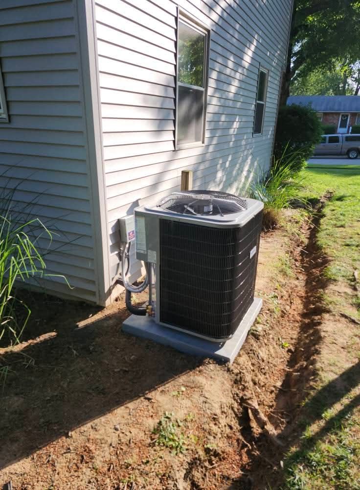 Air conditioner unit outside a light-colored building, sitting on a concrete pad, next to a trench in dirt.