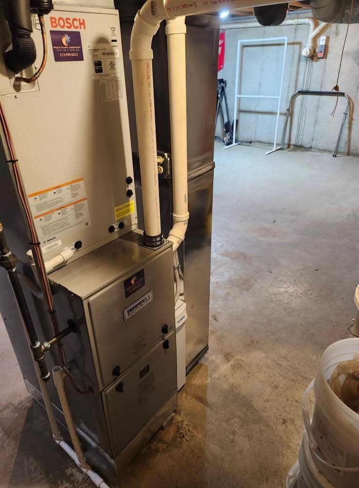 Furnace and air handler unit in a basement. White and silver metal appliances with PVC piping. Concrete floor.