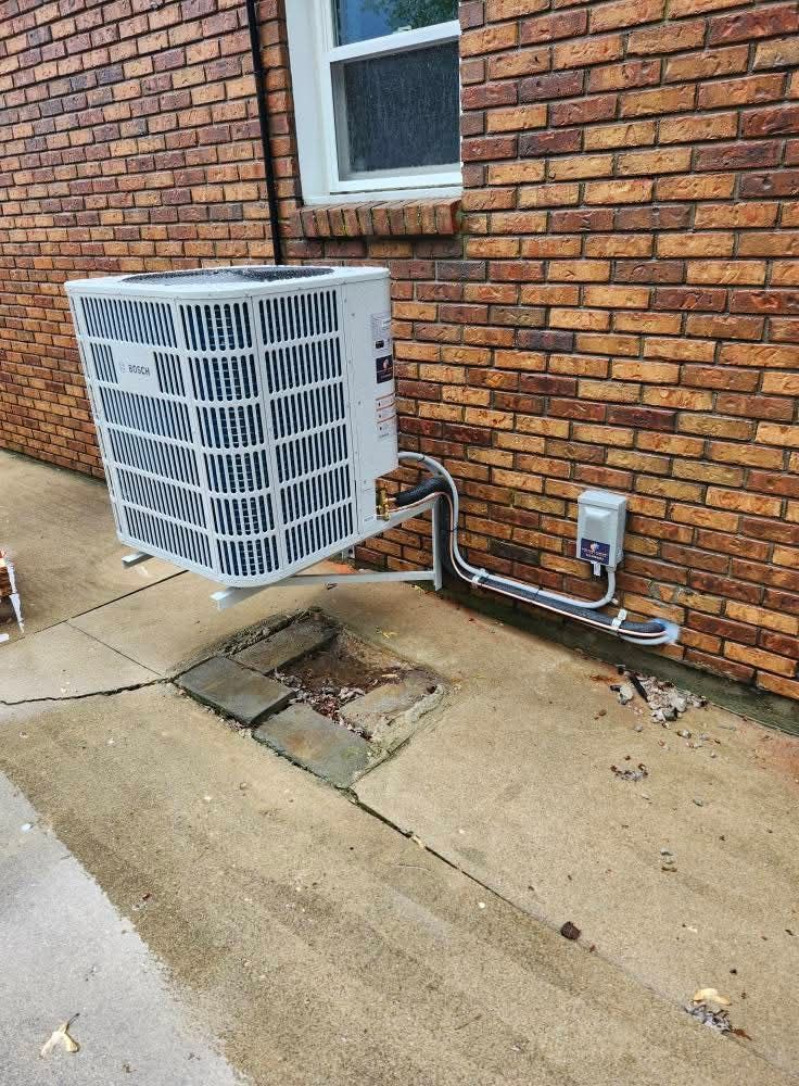 Exterior air conditioning unit mounted on brick wall. Metal pipes connect to electrical box and concrete.