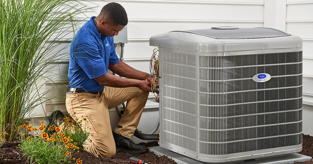 HVAC technician kneels by an air conditioner, working on the unit outside a house.