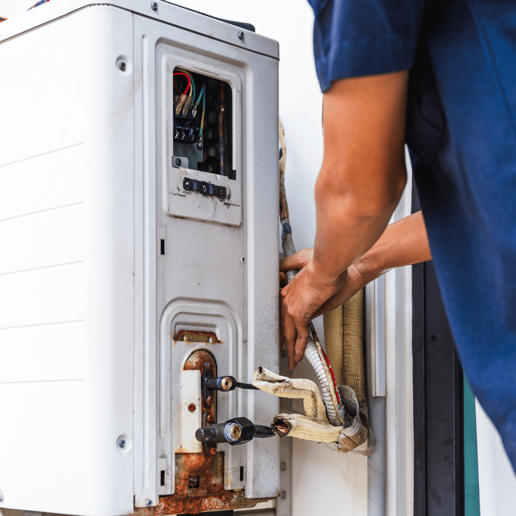 Person in blue shirt repairs a rusty, white outdoor air conditioning unit.