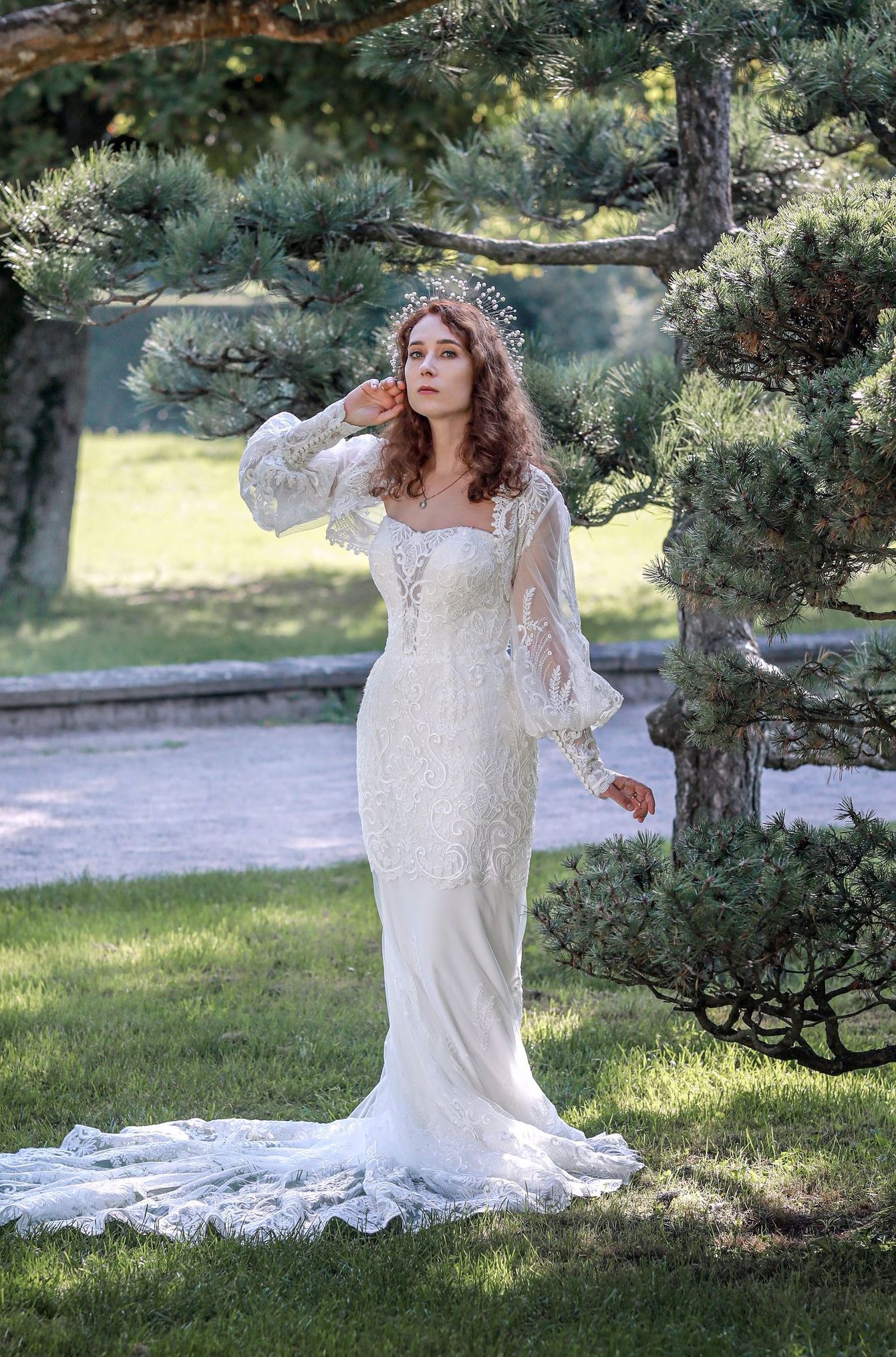 Eine Frau in einem Brautkleid steht im Gras vor einem Baum.