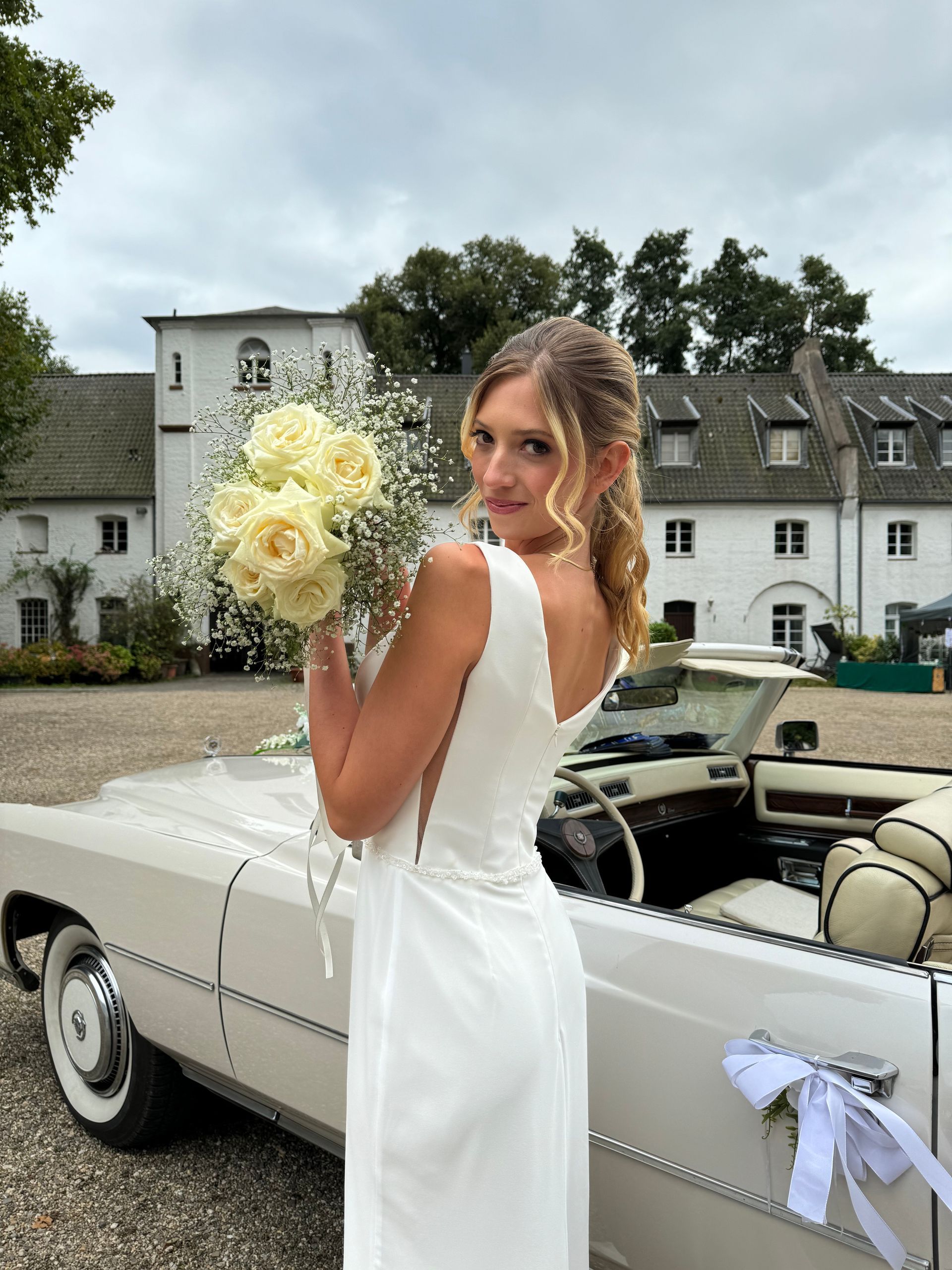 Eine Frau in einem Brautkleid steht neben einem weißen Auto und hält einen Blumenstrauß in der Hand.