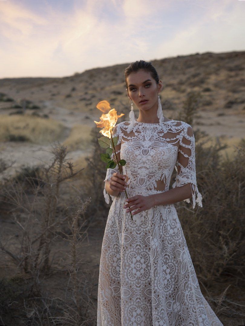 Eine Frau in einem weißen Spitzenkleid hält in der Wüste eine Blume.