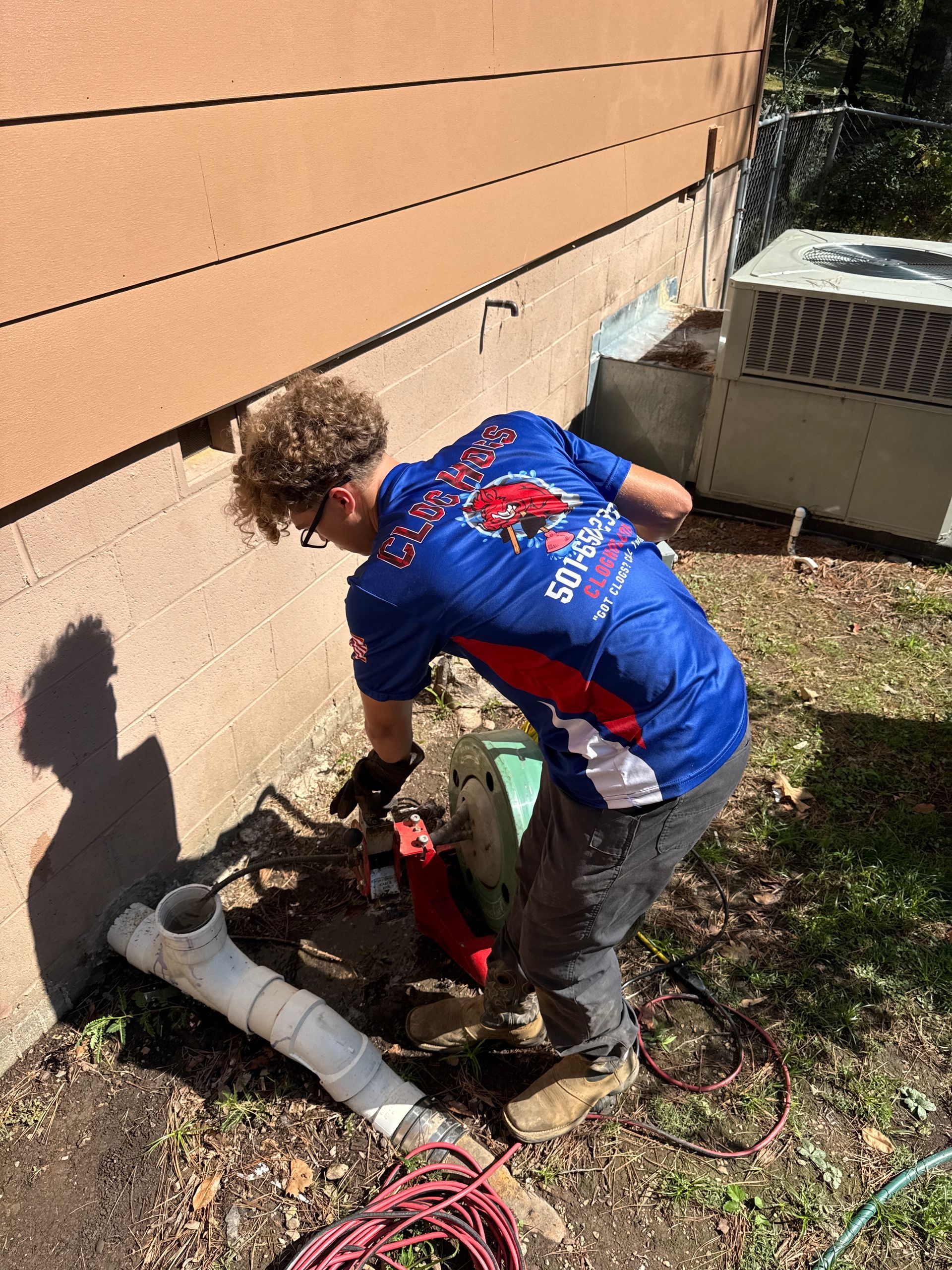 Man in blue shirt using a saw on a pipe near a building, outdoors.