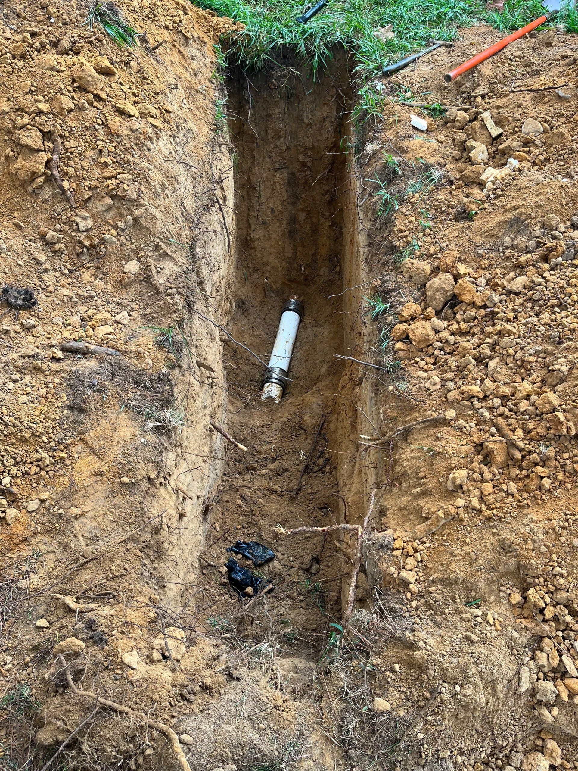 Trench in dirt reveals buried plumbing pipe. Brown soil, green grass.