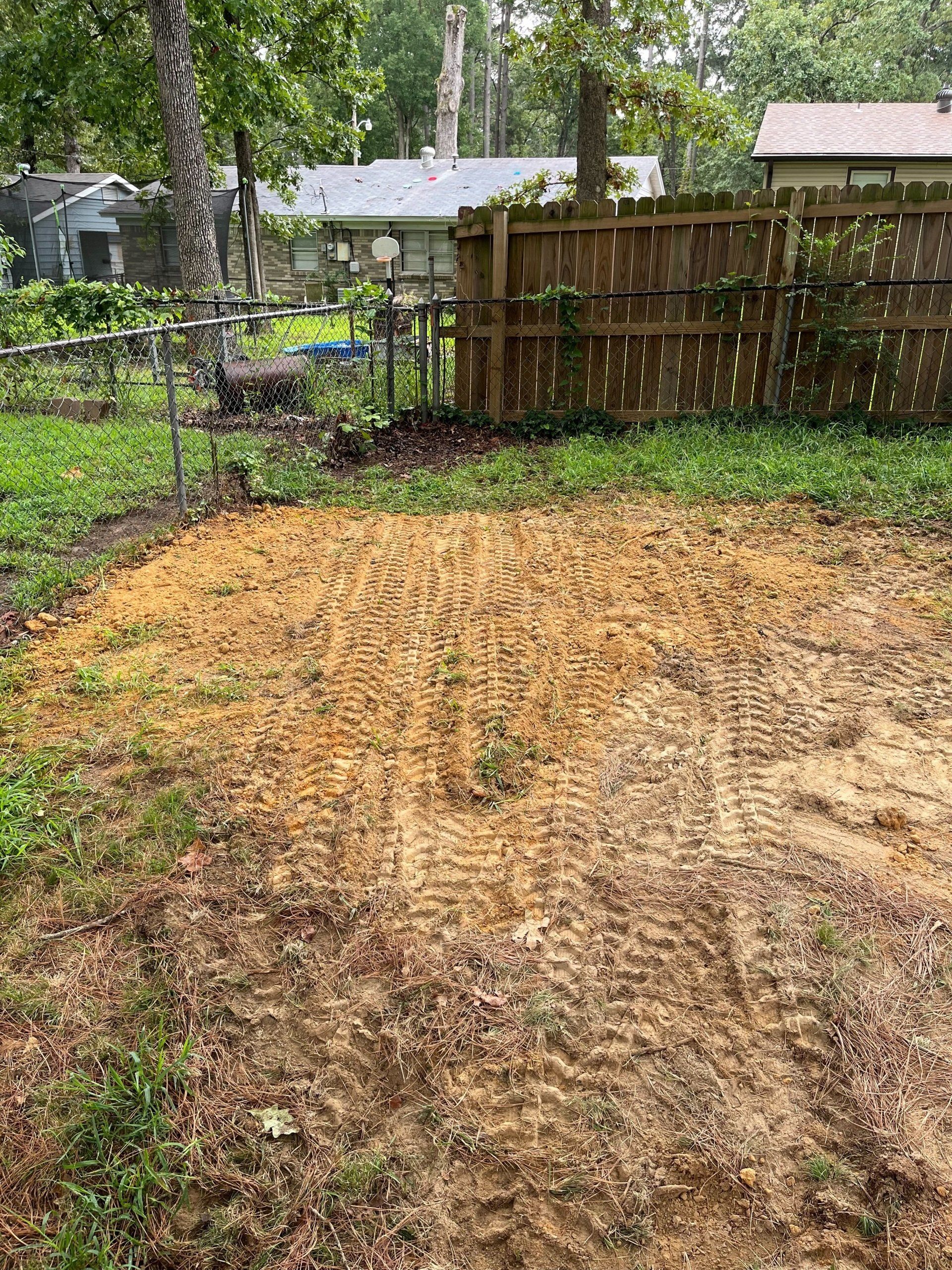 A cleared area of tan soil in a backyard, surrounded by grass and a wooden fence.