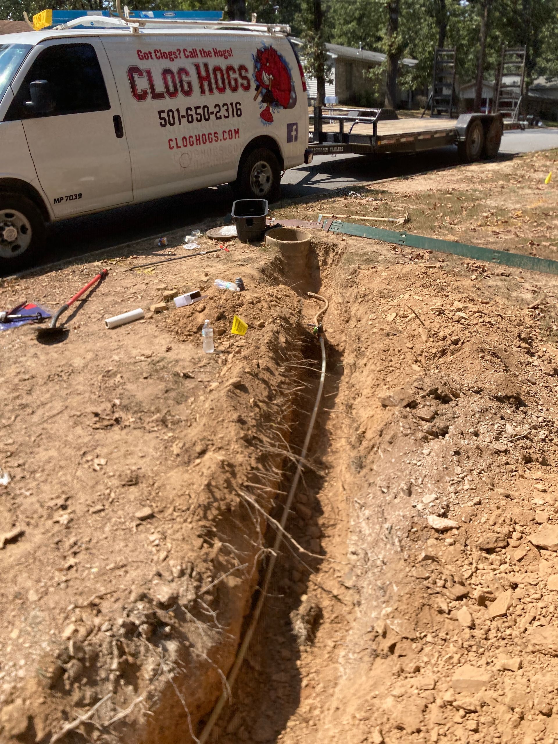 Trench dug with pipe inside; Clog Hogs van parked nearby, tools laid around.
