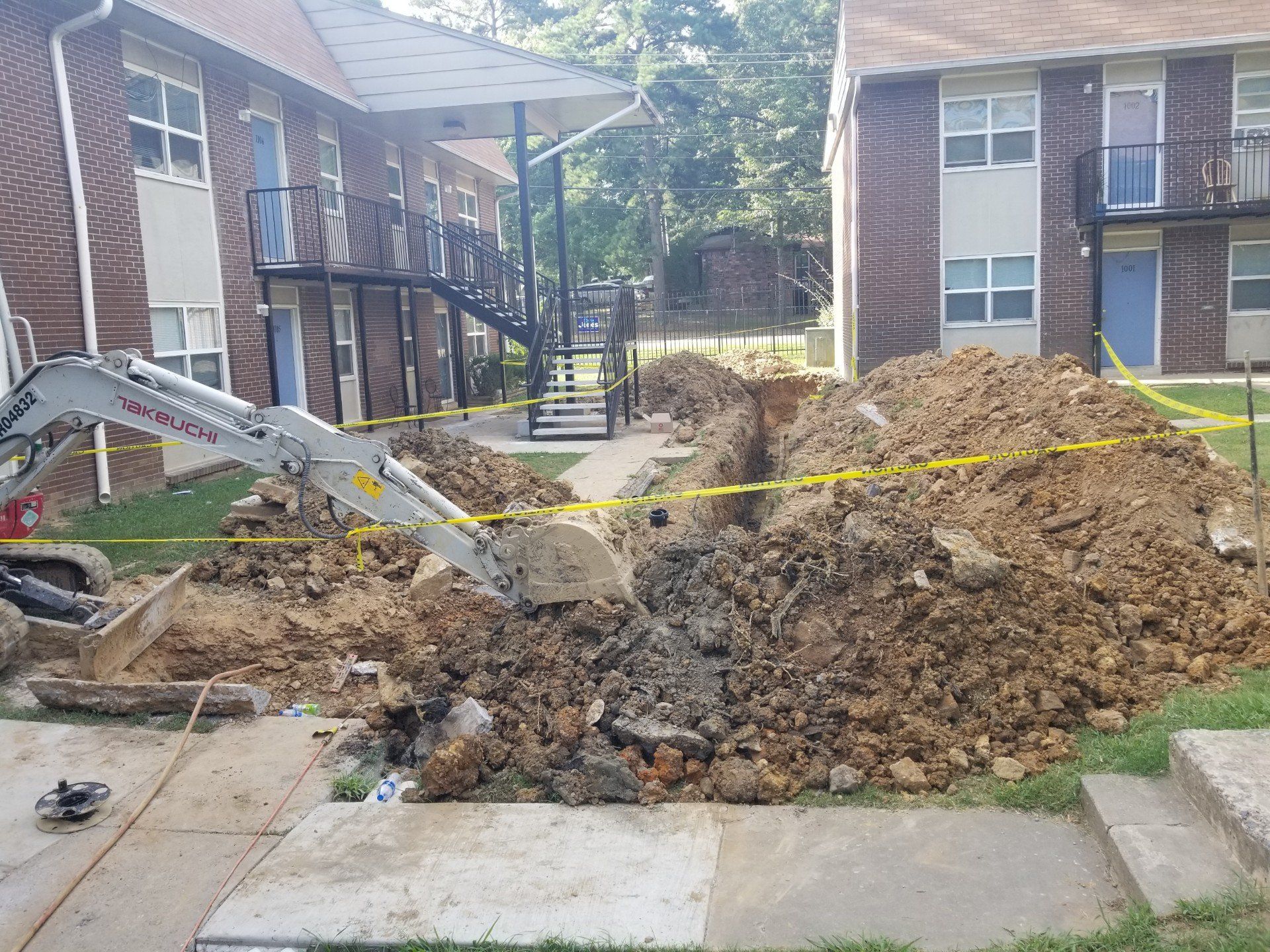 Excavator digging trench in front of apartment buildings. Yellow caution tape and dirt piles are visible.