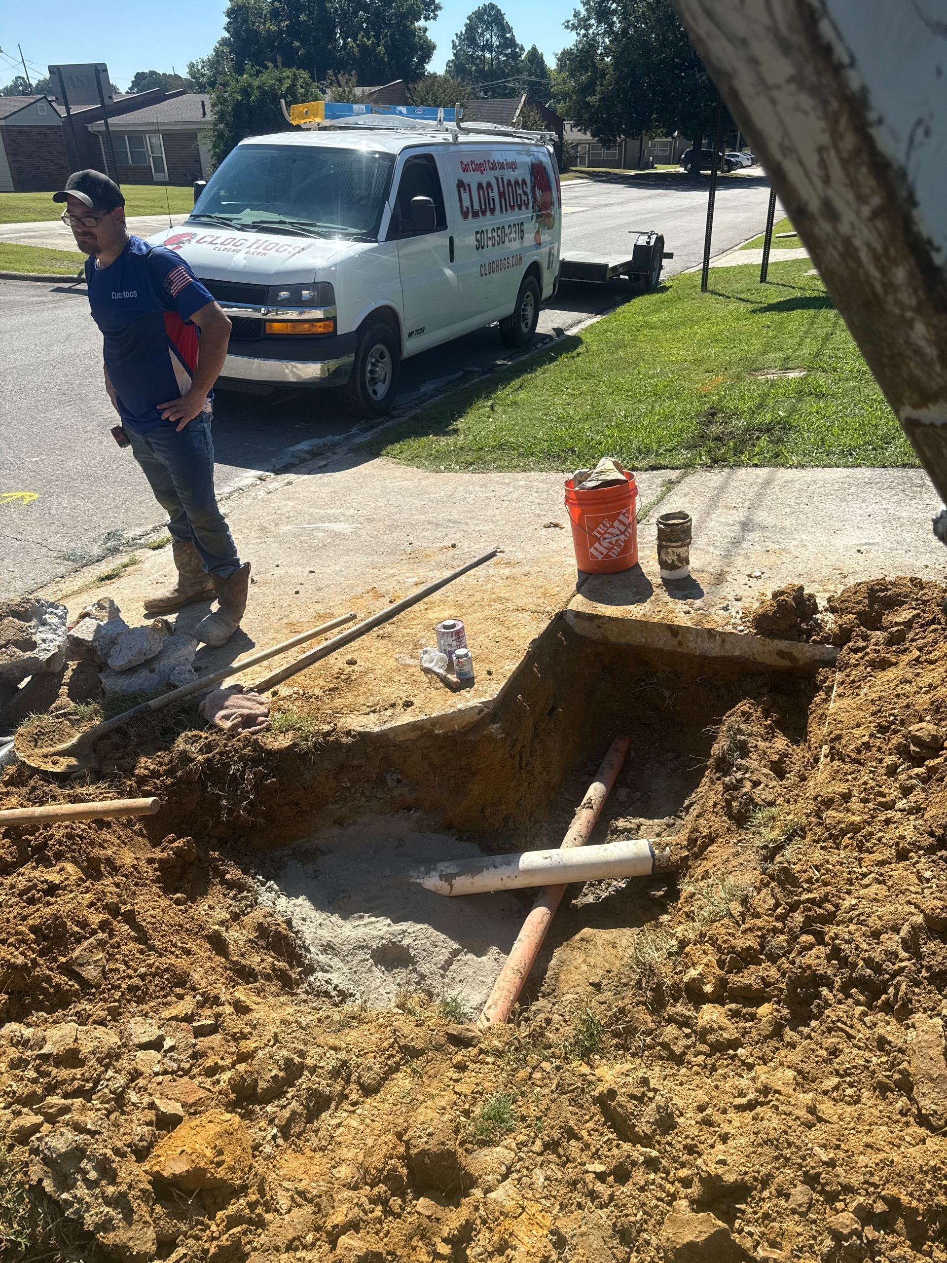 Man by a hole with pipes, next to a work van, a bucket, and other tools.