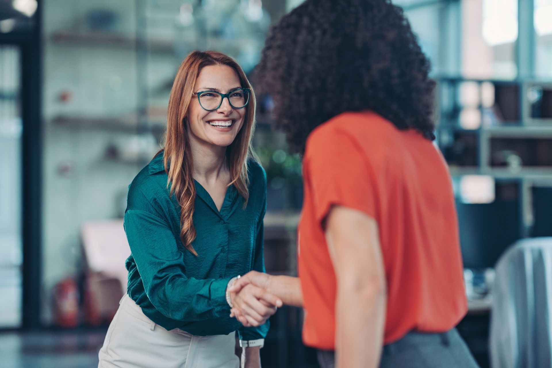 Woman in teal shirt shaking hands with another person in an office setting. Smiling.