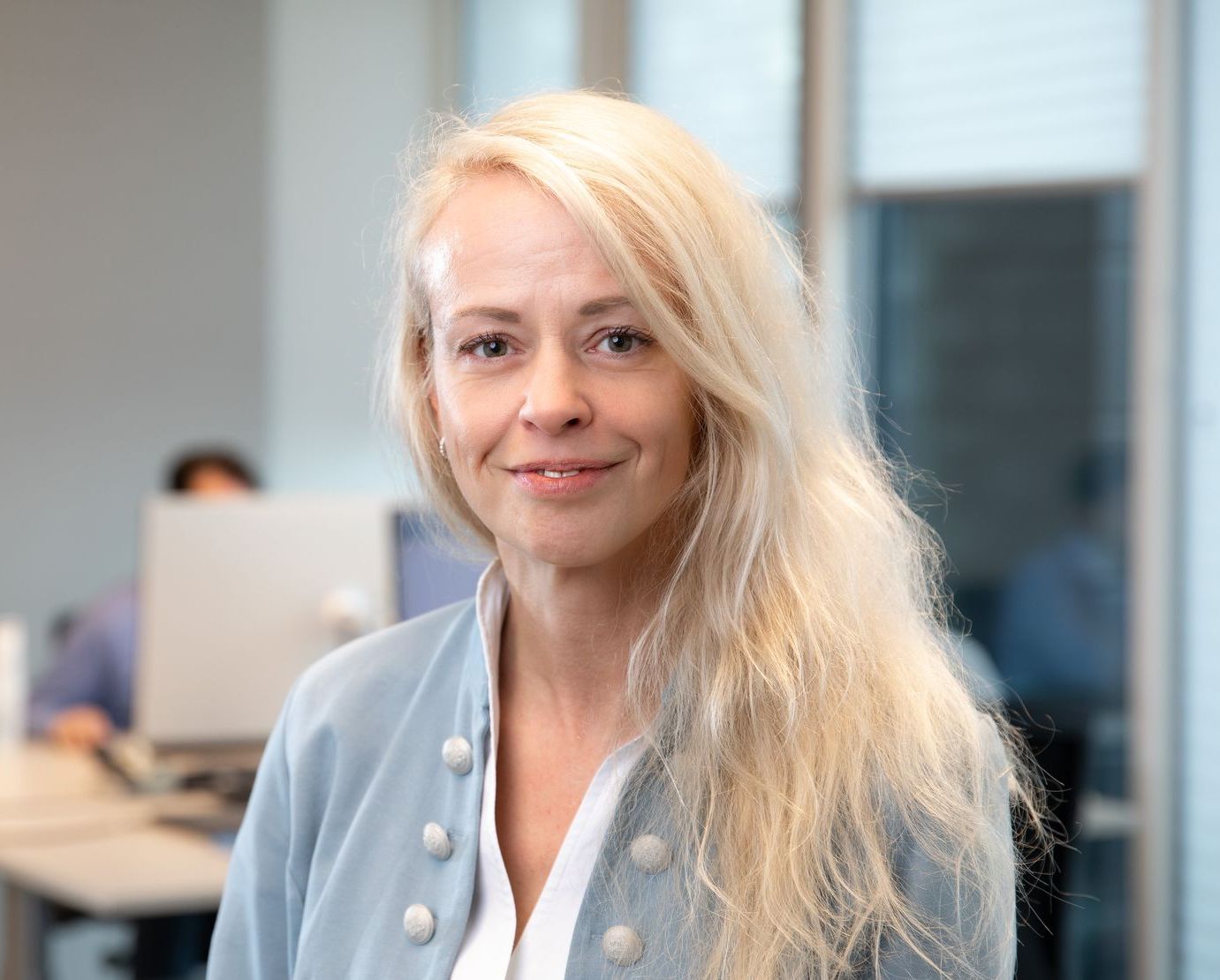 Een vrouw met lang blond haar lacht voor de camera op kantoor.