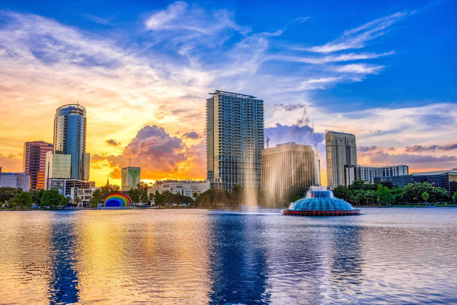 A fountain in the middle of a lake with a city skyline in the background at sunset.