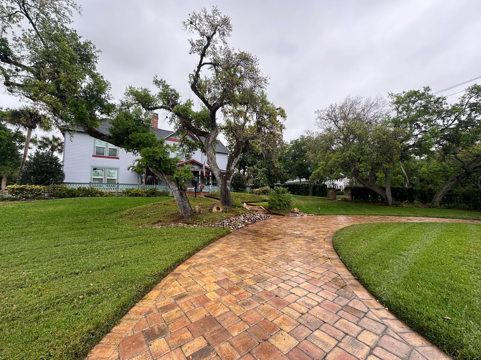 A brick walkway leading to a large white house surrounded by trees.