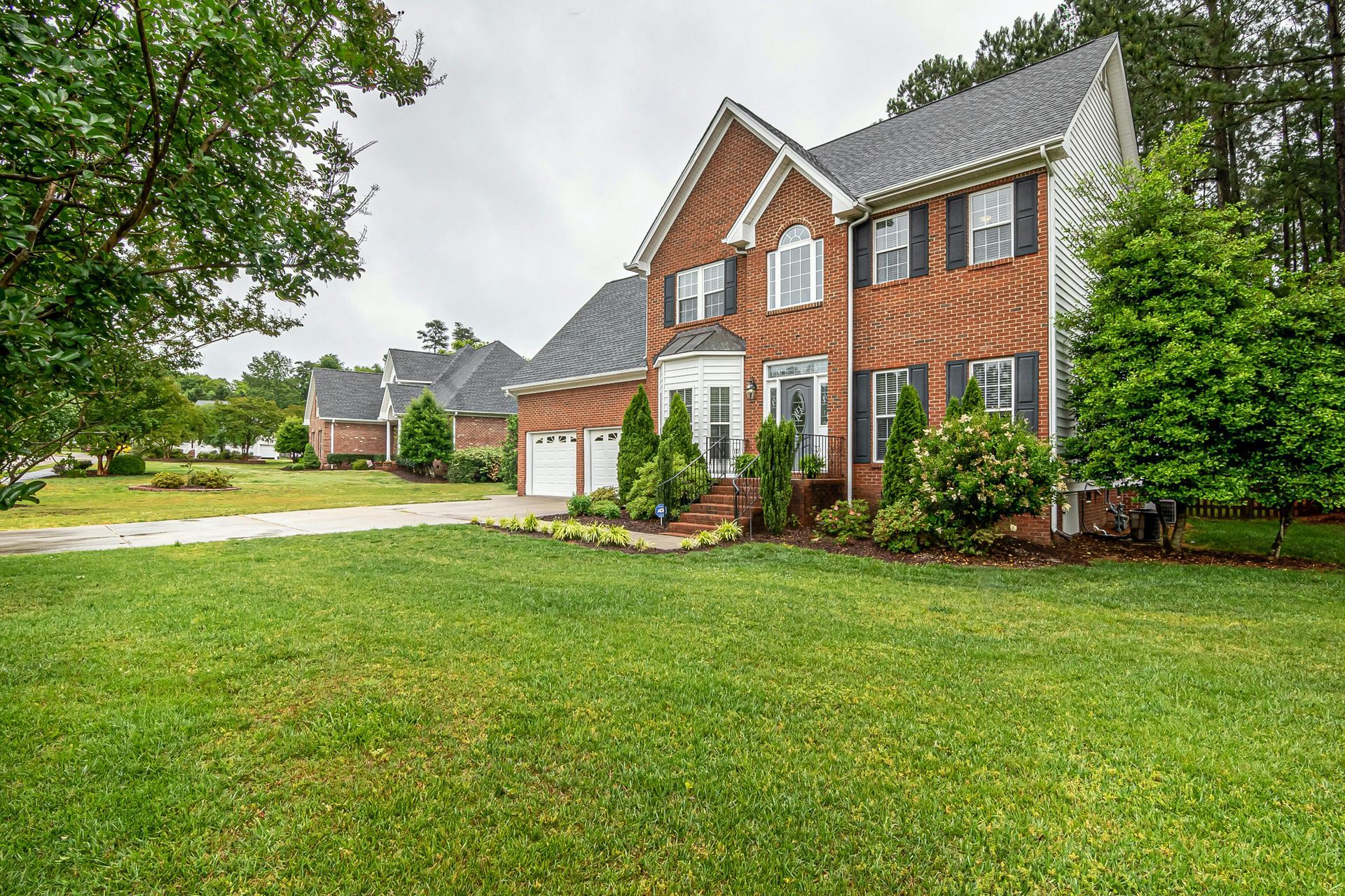 Two-story brick house with dark shutters, front lawn, and trees. Overcast sky.