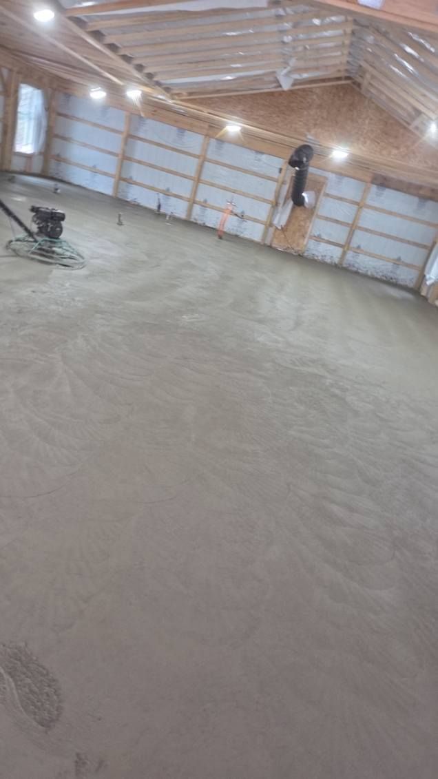 Freshly poured concrete floor being smoothed in a large building with exposed beams and overhead lights.