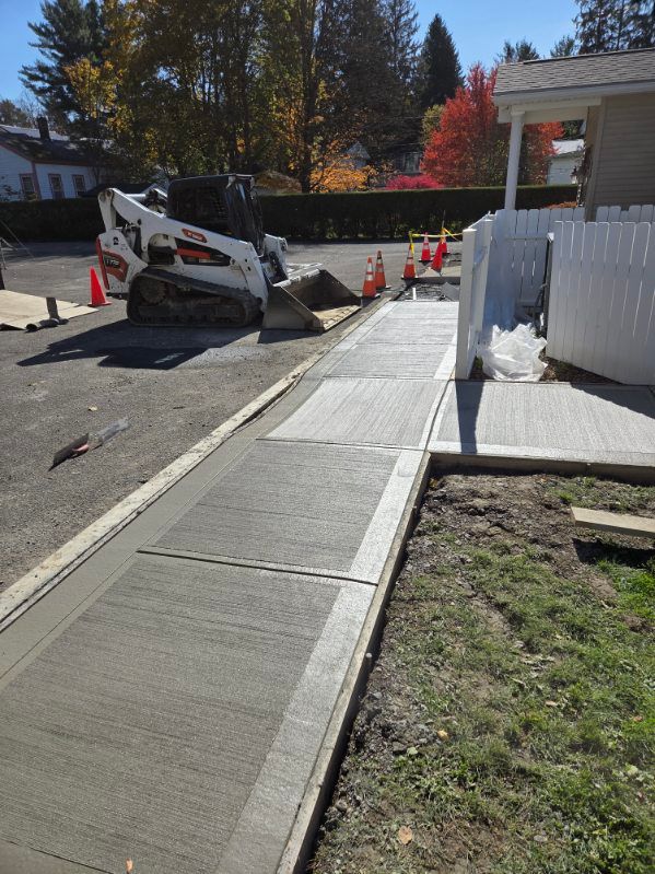 Concrete sidewalk construction site with a skid steer loader and orange traffic cones.