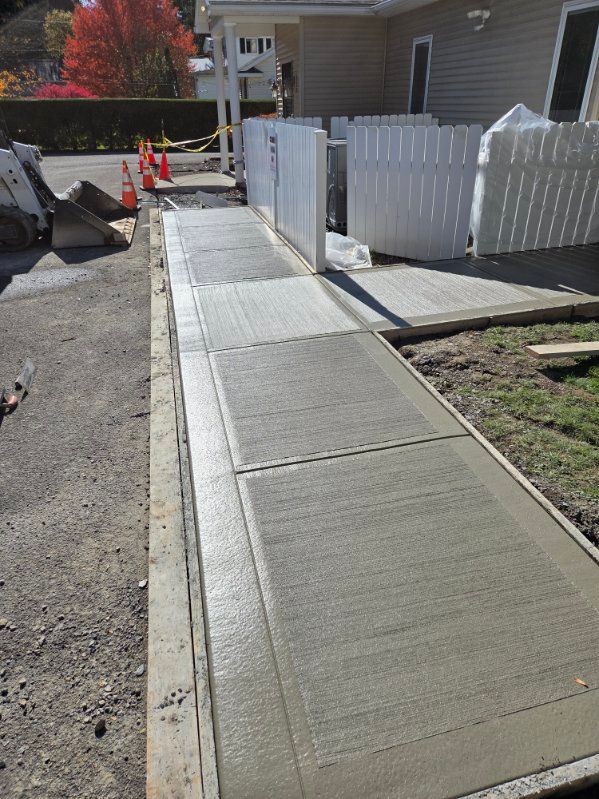 Freshly poured concrete sidewalk next to a building, fenced area, construction cones visible.