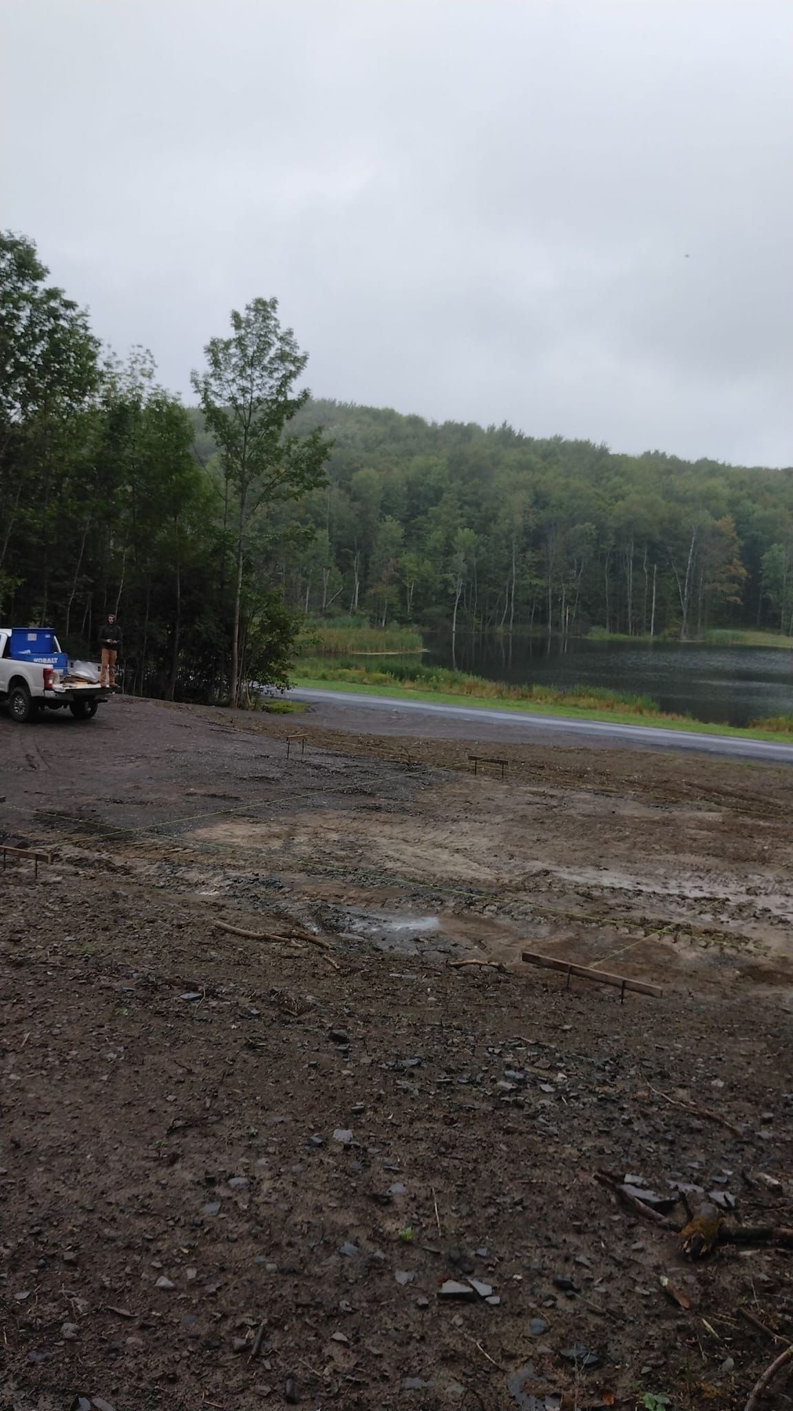 A white truck parked on a gravel lot next to a body of water and a forested hill under a cloudy, overcast sky.