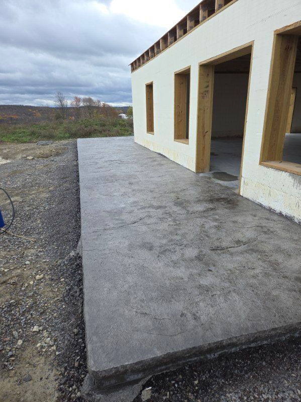 Concrete patio next to unfinished building. Gravel border, overcast sky in background.
