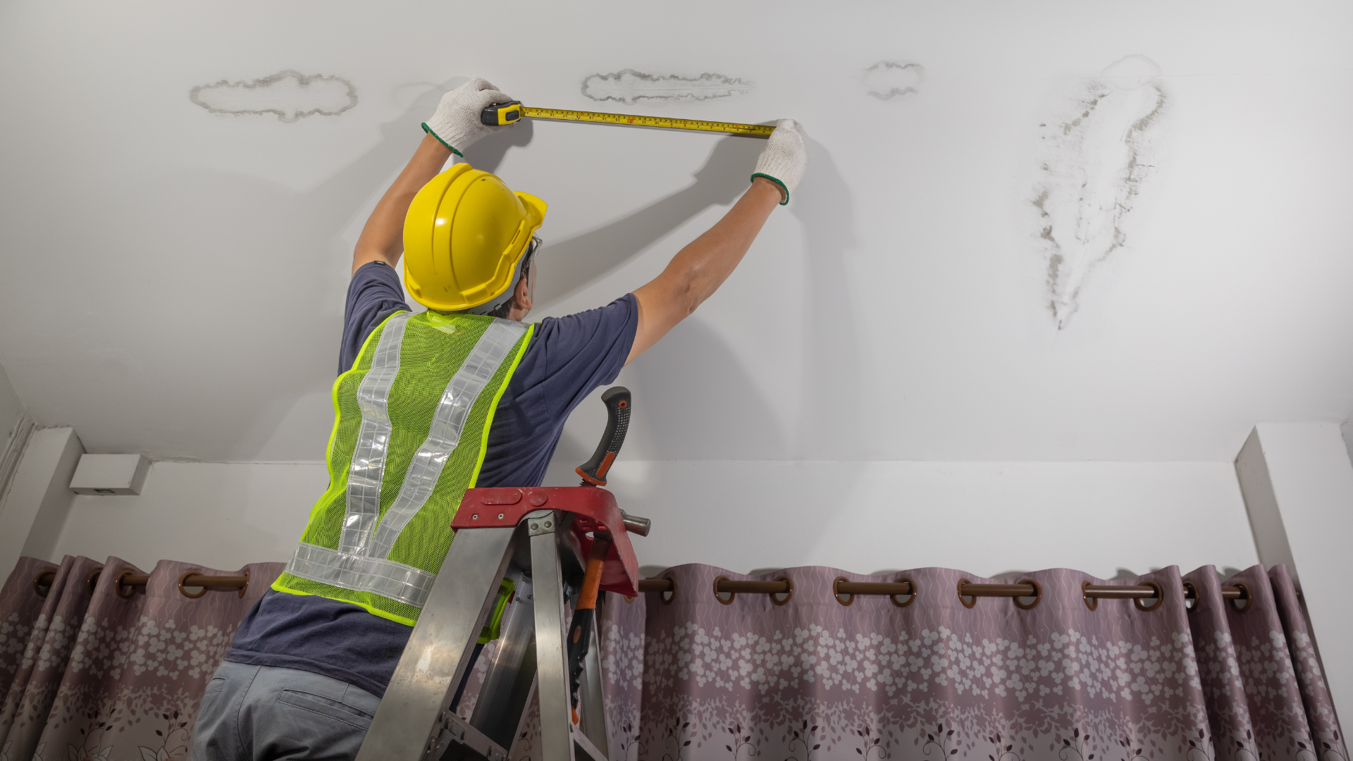A man is measuring a wall with a tape measure while sitting on a ladder.