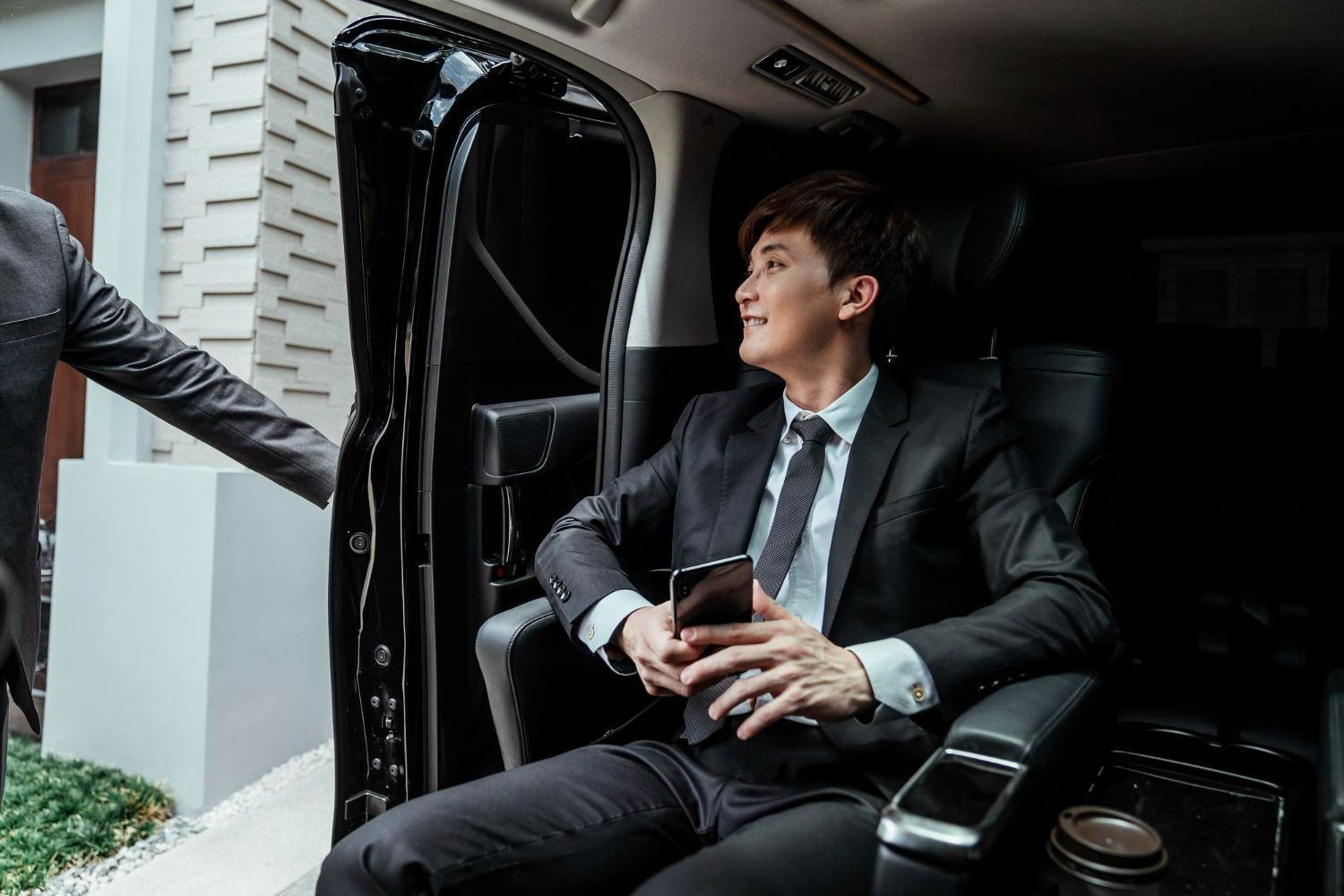 Man in suit seated in a black car, looking to the right, holding a phone. A person is opening the car door.
