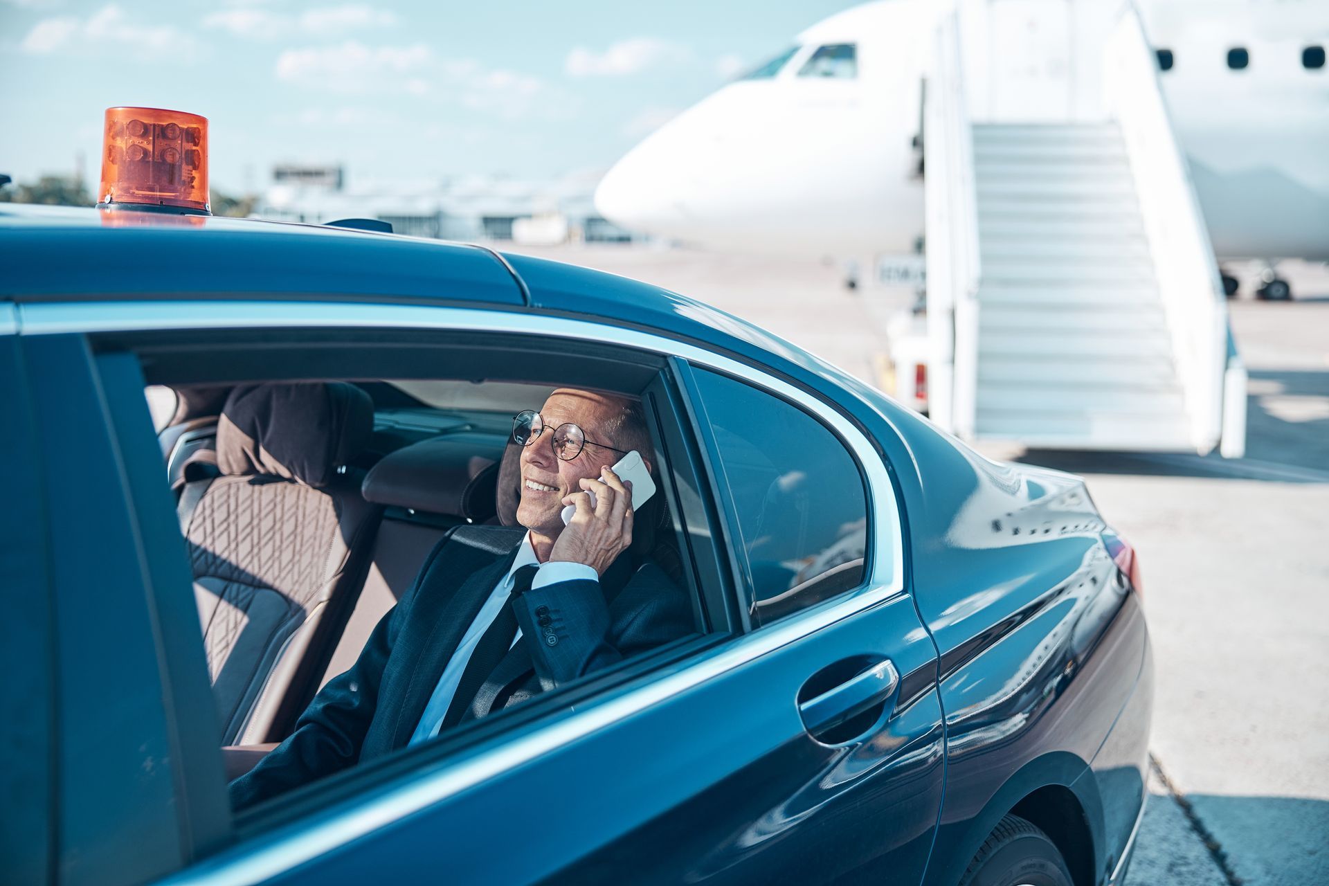 Suit-clad passenger in a blue car talking on a phone beside a private jet runway