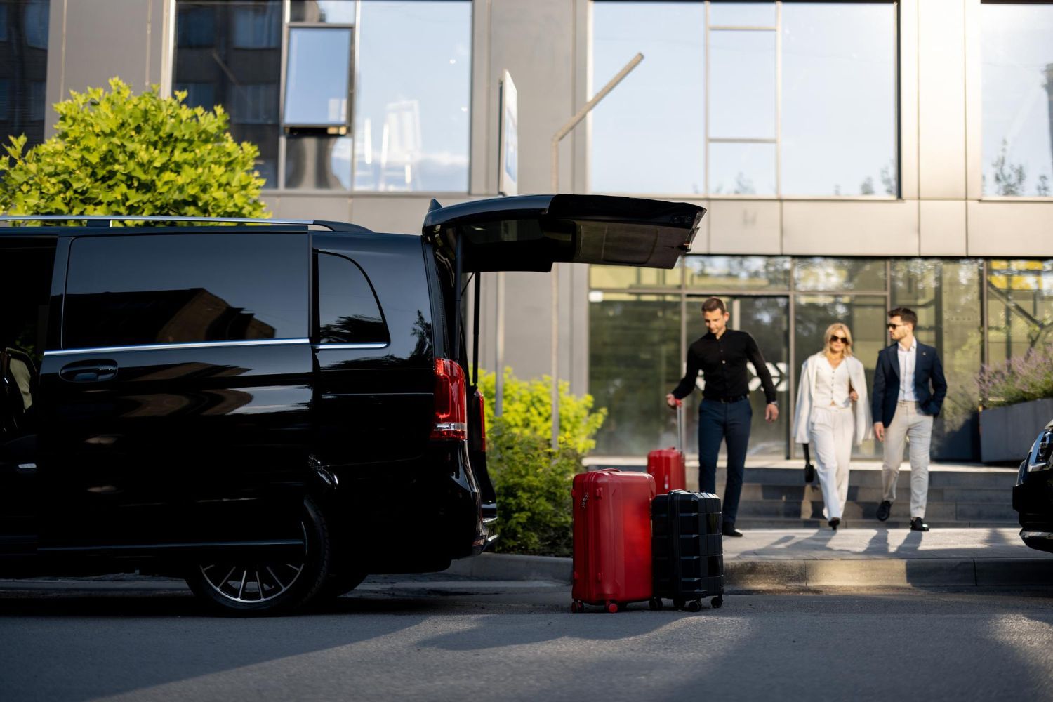 Black van with open trunk, two people exiting building, suitcases nearby.