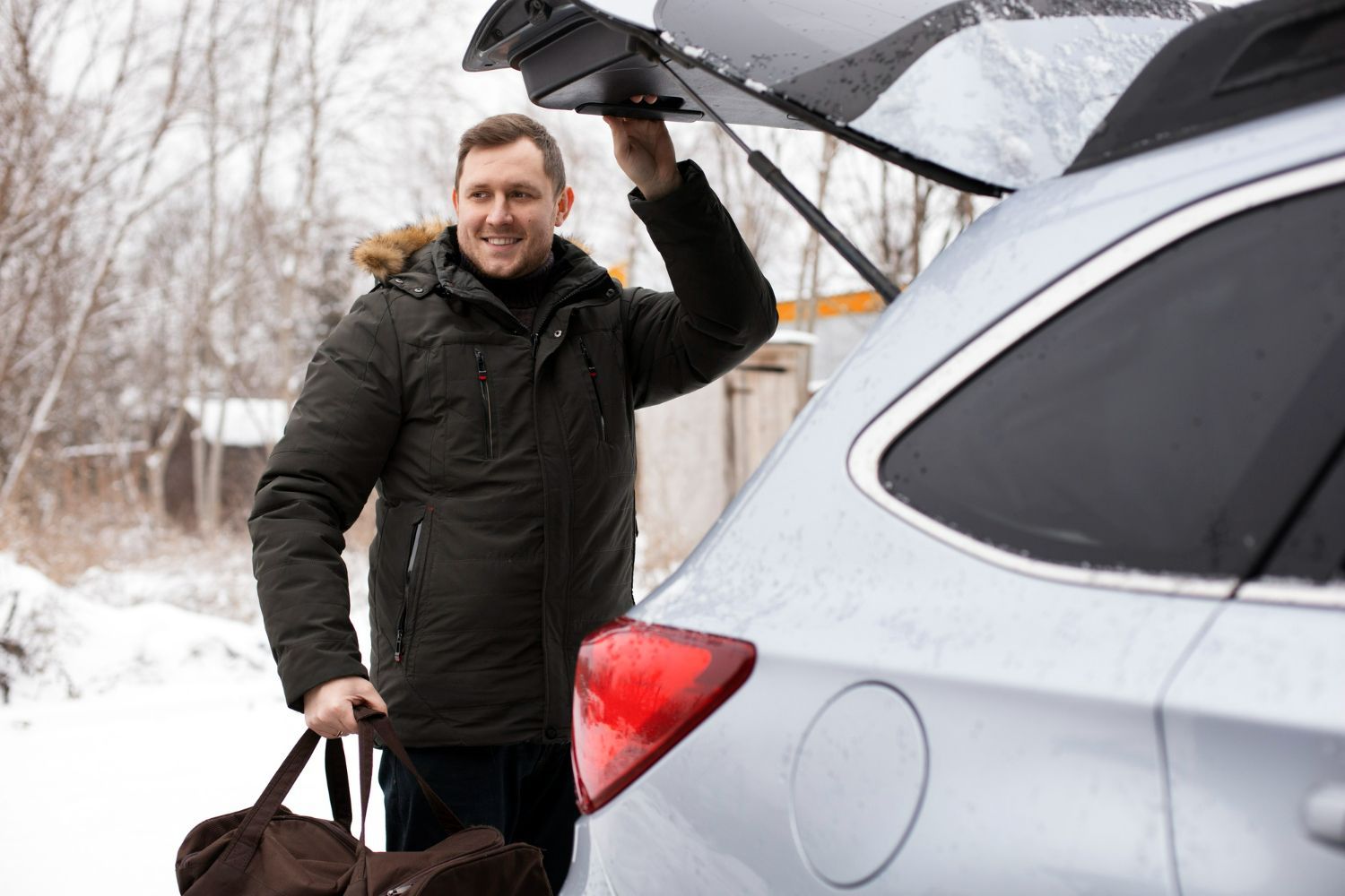 Man opening car trunk, holding bag, smiling outside in a snowy environment.