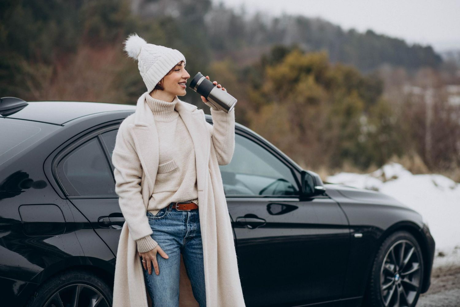 Woman in winter wear drinks from a thermos next to a black car in a snowy landscape.