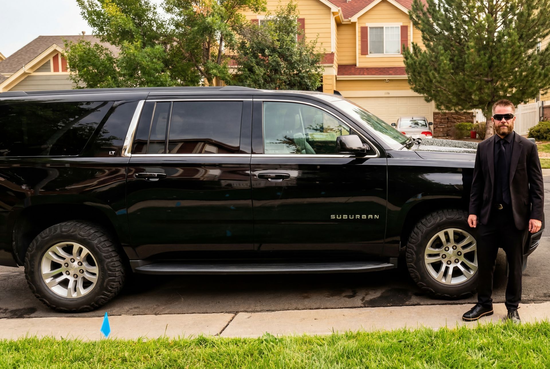 Man in black suit stands beside a black SUV on a suburban street.