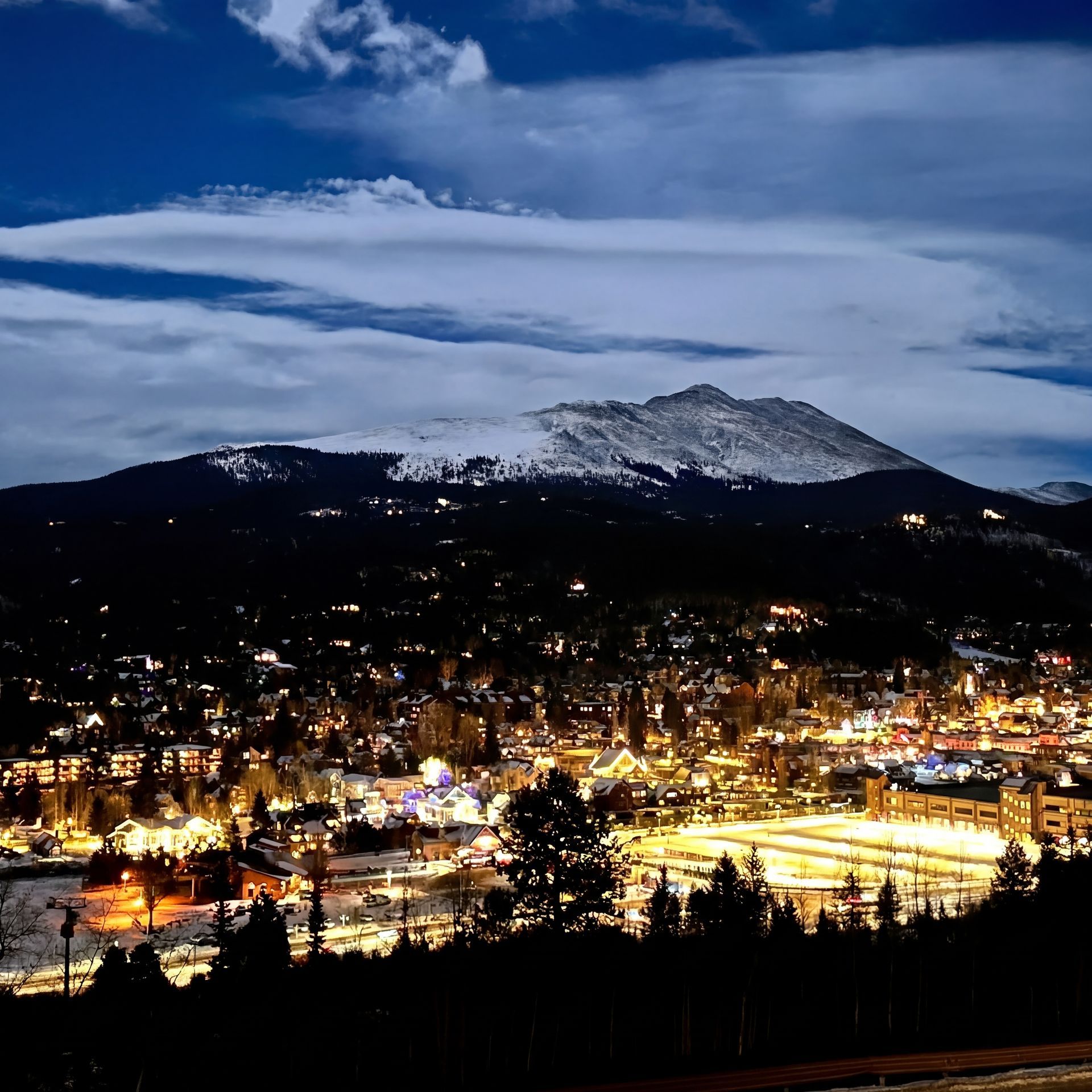 Town at night, snow-capped mountain backdrop. City lights illuminate buildings; dark trees in foreground, blue sky.