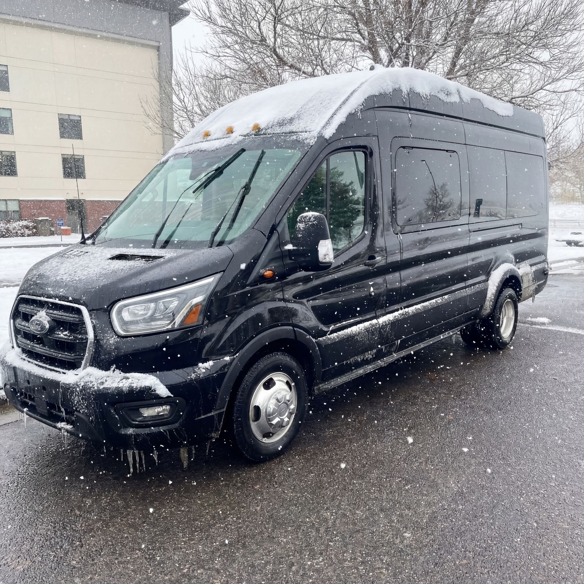 Black passenger van covered in snow parked on asphalt during a snowfall.