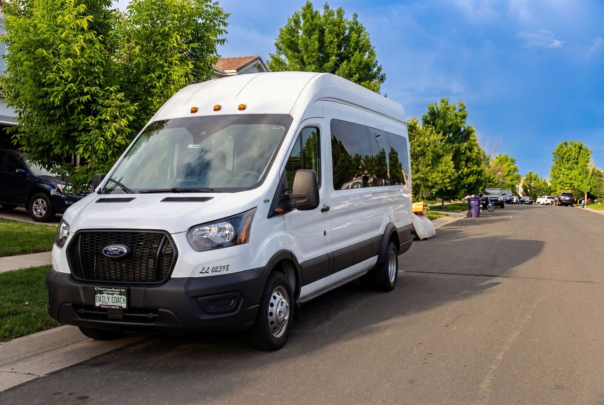 White passenger van parked on a residential street; trees, houses, and blue sky visible.