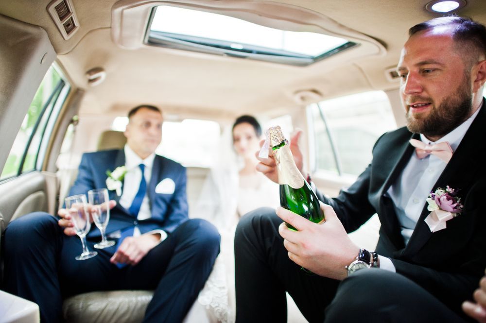 A bride and groom are sitting in a limousine with a man holding a bottle of champagne.