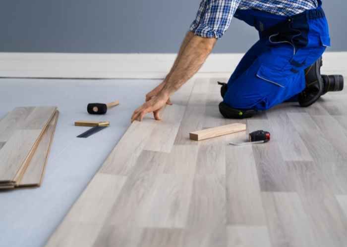 A man is installing a new wooden floor in a room with large windows, focusing on the details of the installation process.