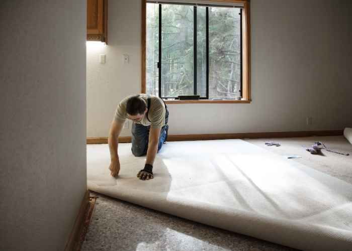 A person is carefully removing a section of carpet near a wall baseboard.
