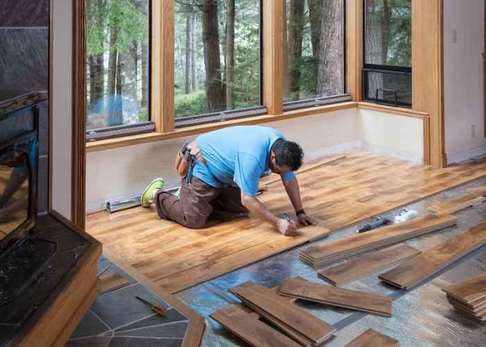 A man is installing a new wooden floor in a room with large windows, focusing on the details of the installation process.