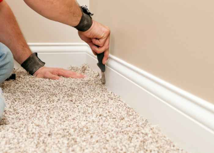 A person is carefully removing a section of carpet near a wall baseboard.