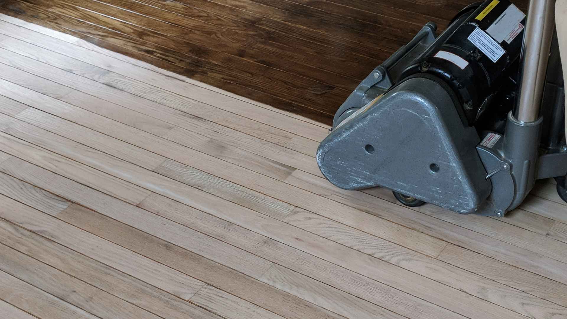 A floor sander is positioned on a light-colored wooden floor, with a section of dark-stained wood beside it.