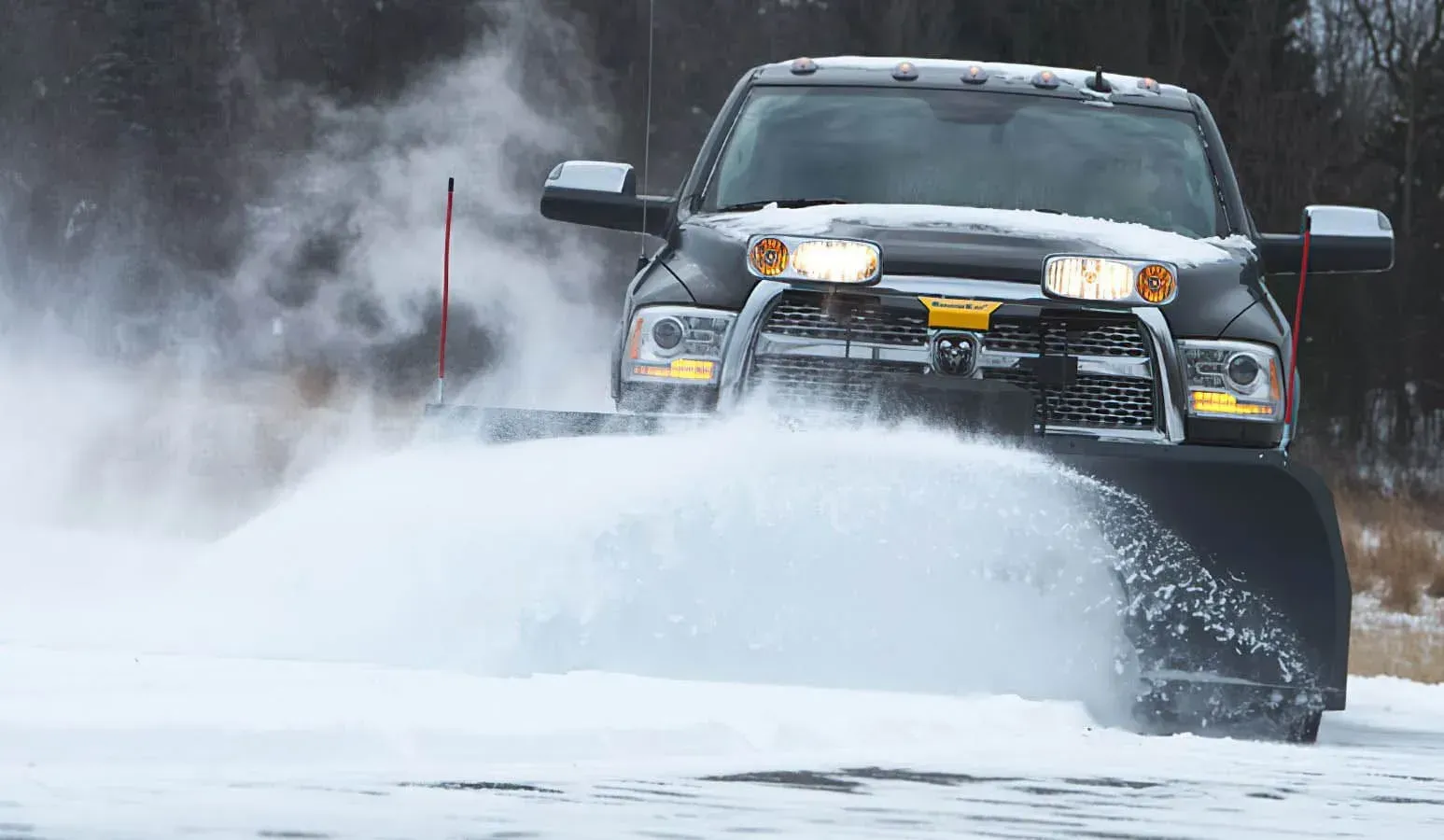 Black snowplow truck clearing snow on a road, white snow spraying forward.