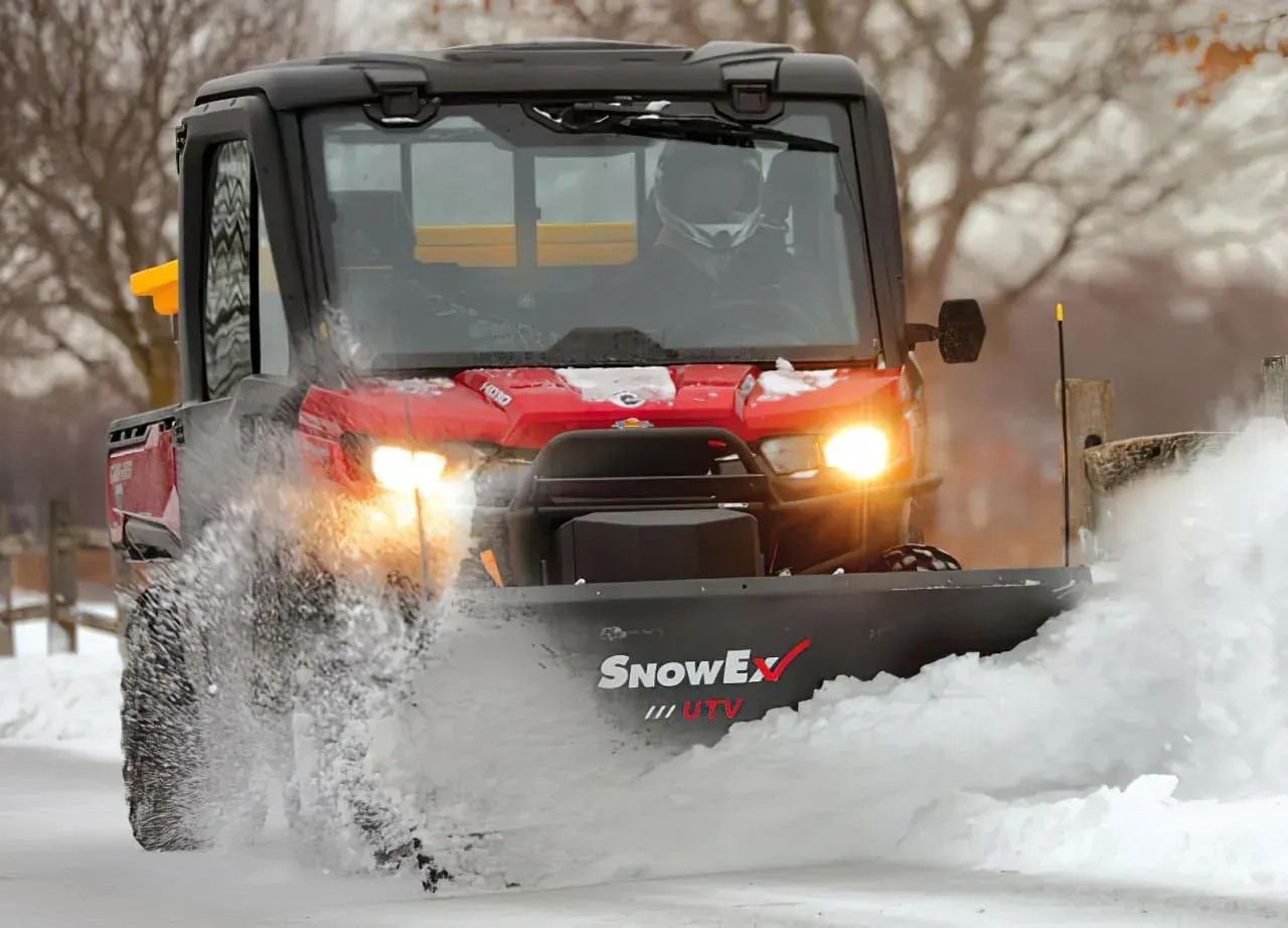 Red snowplow truck plowing snow on a road, with headlights on.