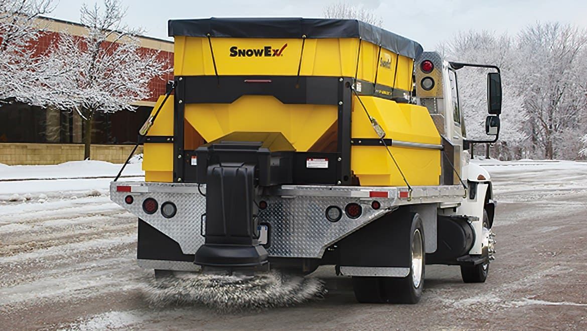 Yellow snowplow truck spreading salt on a snow-covered road.