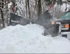 A truck with a snow plow pushing a large pile of snow in a snowy, wooded area.