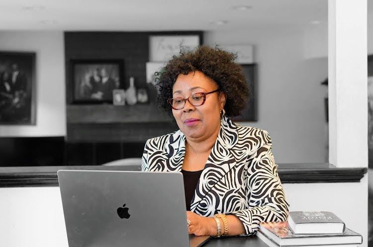Woman wearing glasses and blazer, using a laptop, sitting at a table with books.